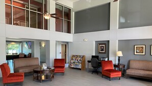 A spacious lobby with a high ceiling, a ceiling fan, and a seating area with red chairs and sofas. There is a desk with a computer and a printer, and a wall-mounted TV. - Ramada by Wyndham Luling (Luling)
