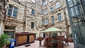 Un patio con una mesa y sillas bajo una pérgola, rodeado por un edificio con muros cubiertos de hiedra y múltiples ventanas.