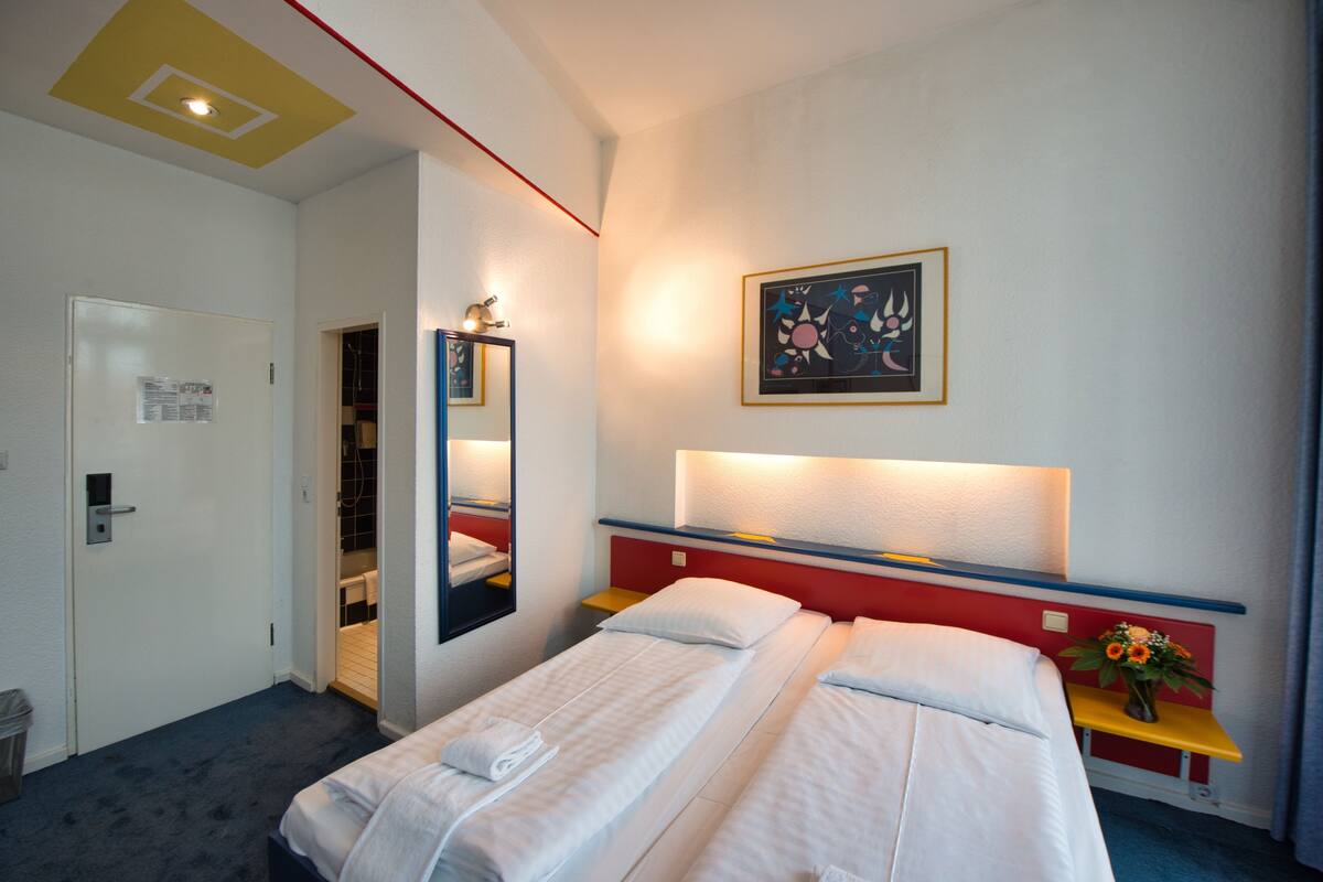Habitación de hotel con dos camas, un espejo, un cuadro y una mesita con un florero.