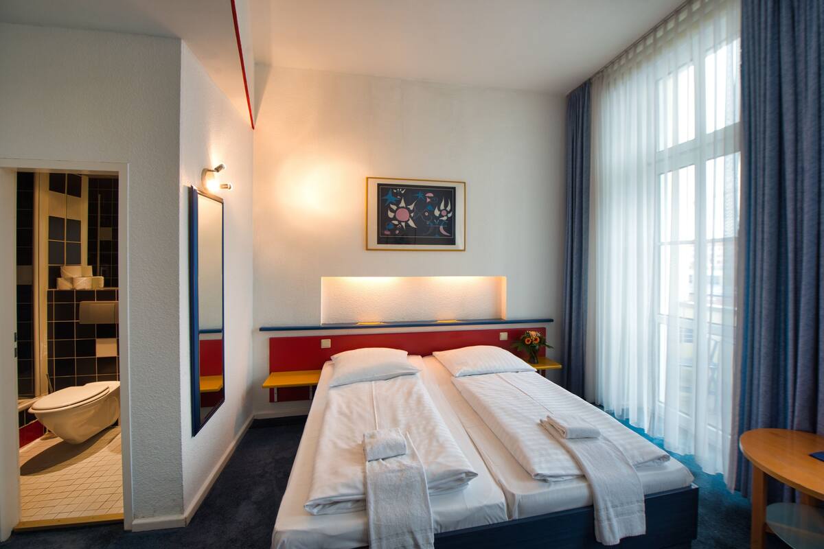 Habitación de hotel con cama, mesita de noche, espejo, ventana con cortinas y baño con inodoro y lavamanos.