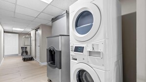 A laundry room with a front-loading washer and dryer, a water dispenser, and a cart. - Days Inn & Suites by Wyndham Peachtree Corners/Norcross (Peachtree Corners)