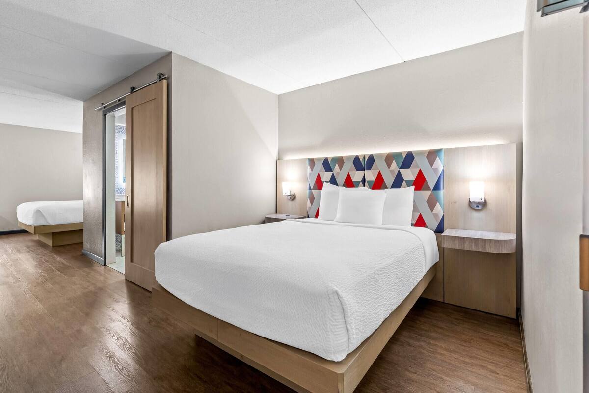 Habitación de hotel moderna con una cama grande, piso de madera, puerta de cristal y cabecera con diseño geométrico.