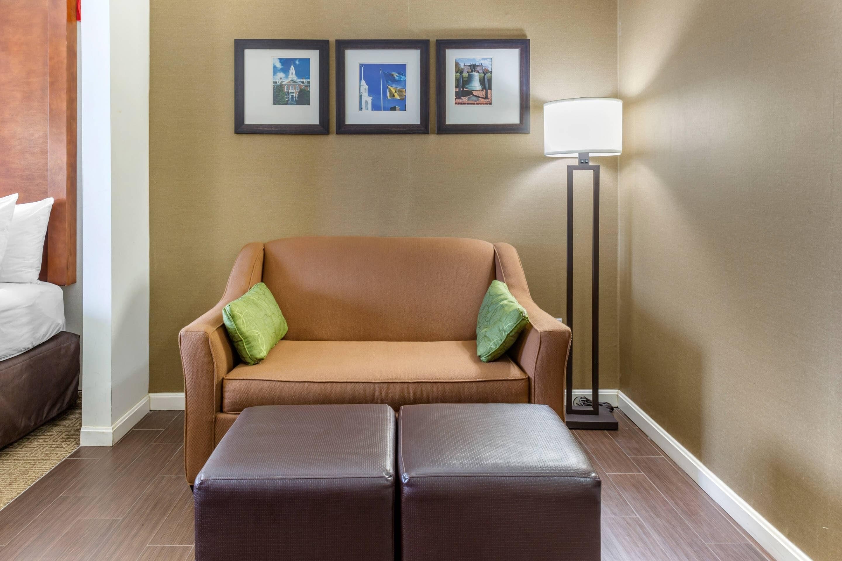 Habitación de hotel con un sofá marrón, dos almohadas verdes, un reposapiés marrón, una lámpara de pie y tres cuadros en la pared.