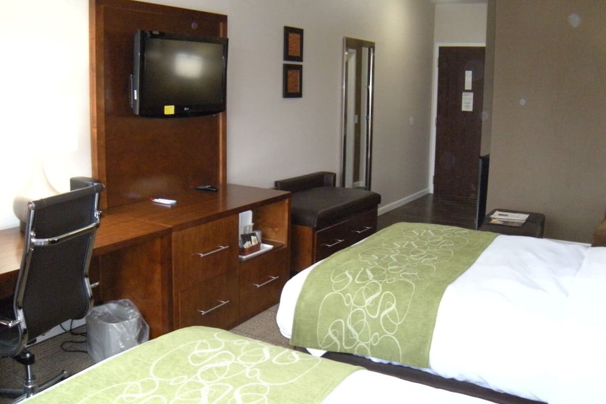 A hotel room with a TV mounted on a wooden cabinet, a desk with a chair, a bed with green bedding, and a trash bin.