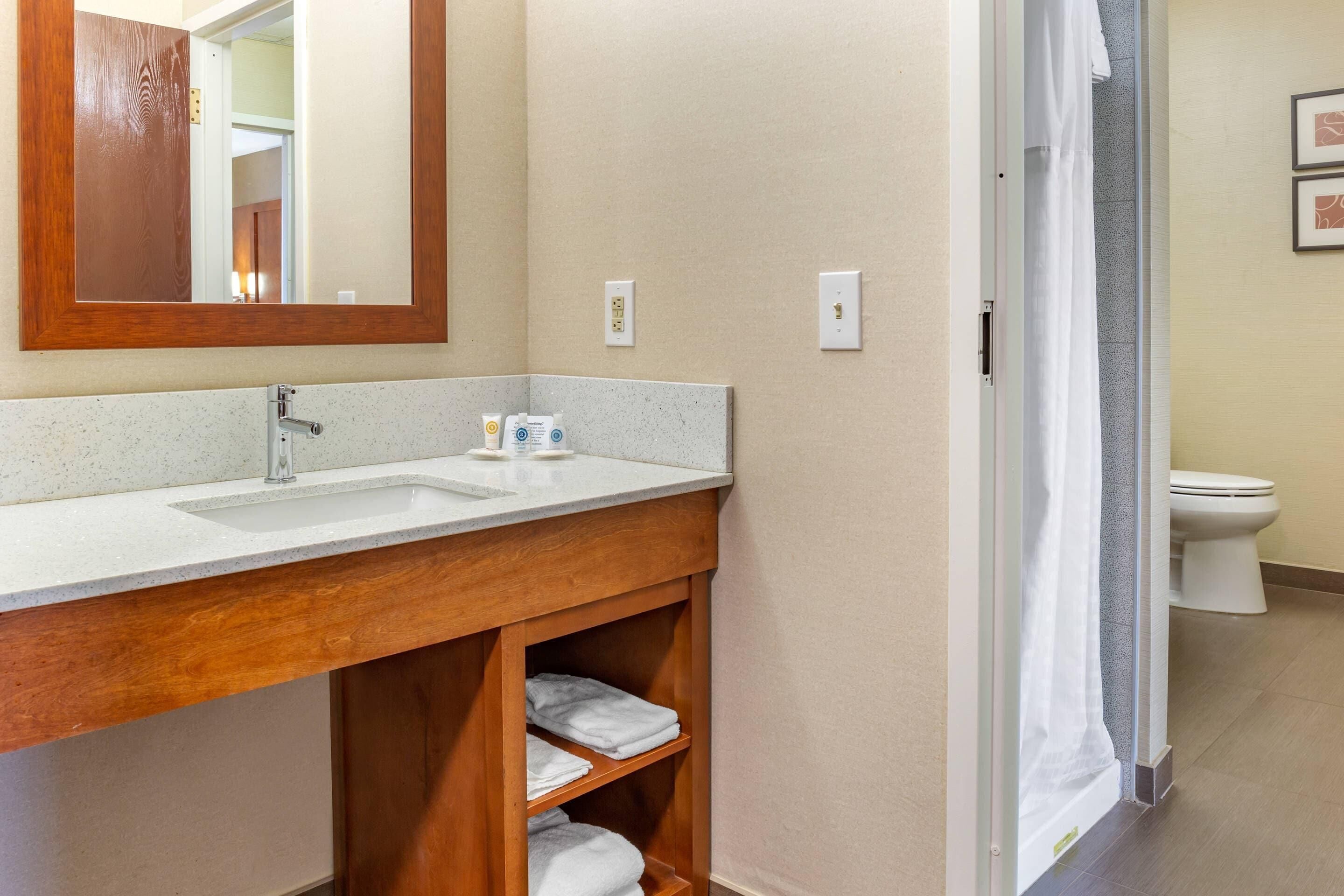 Une salle de bain avec un meuble-lavabo en bois, un évier et un miroir. On y trouve des serviettes sur les étagères et un rideau de douche.