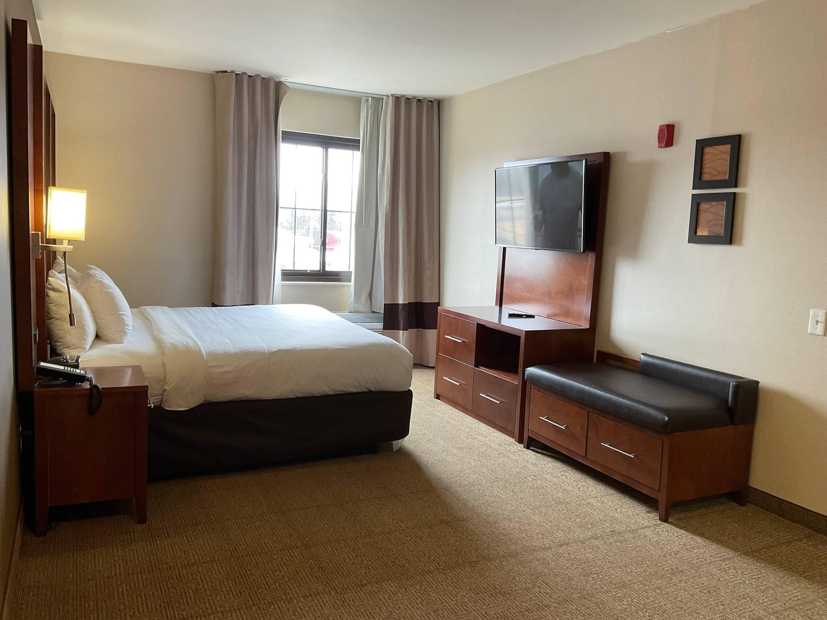 A hotel room with a bed, a TV on a wooden stand, a desk with a lamp, and a window with curtains.