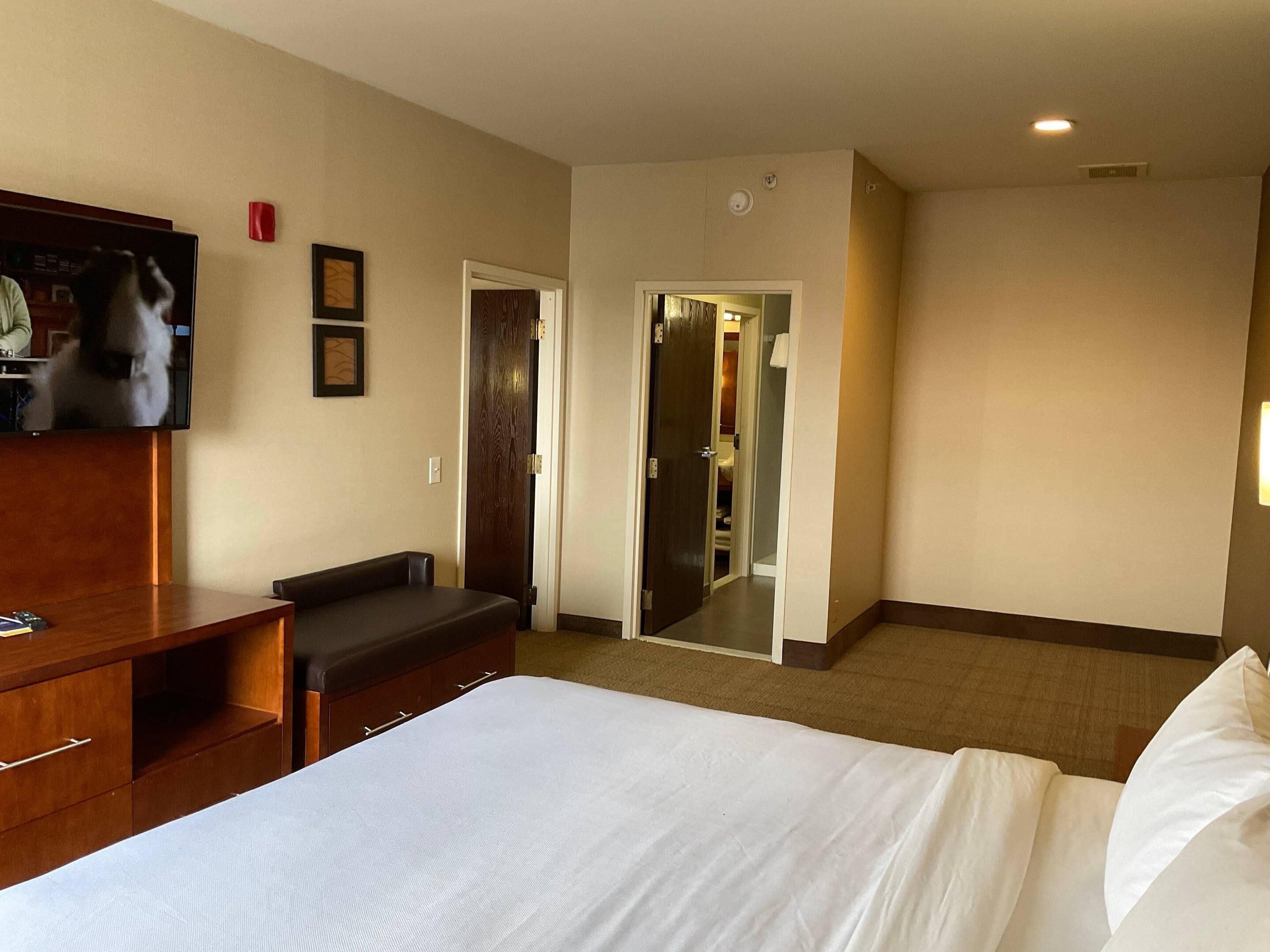 A hotel room with a bed, a wooden bench, a TV, and a door leading to a bathroom.