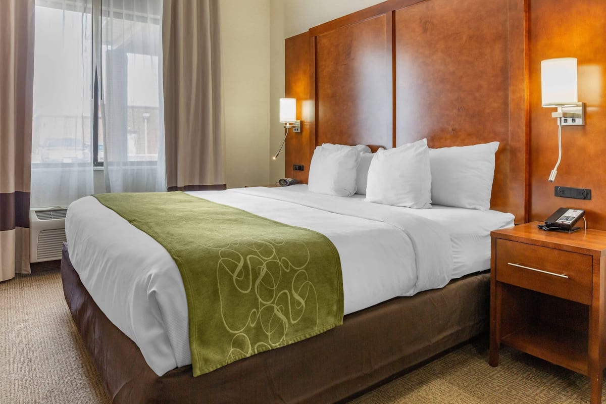A hotel room with a large bed, a wooden headboard, two bedside lamps, a telephone, and a window with curtains.