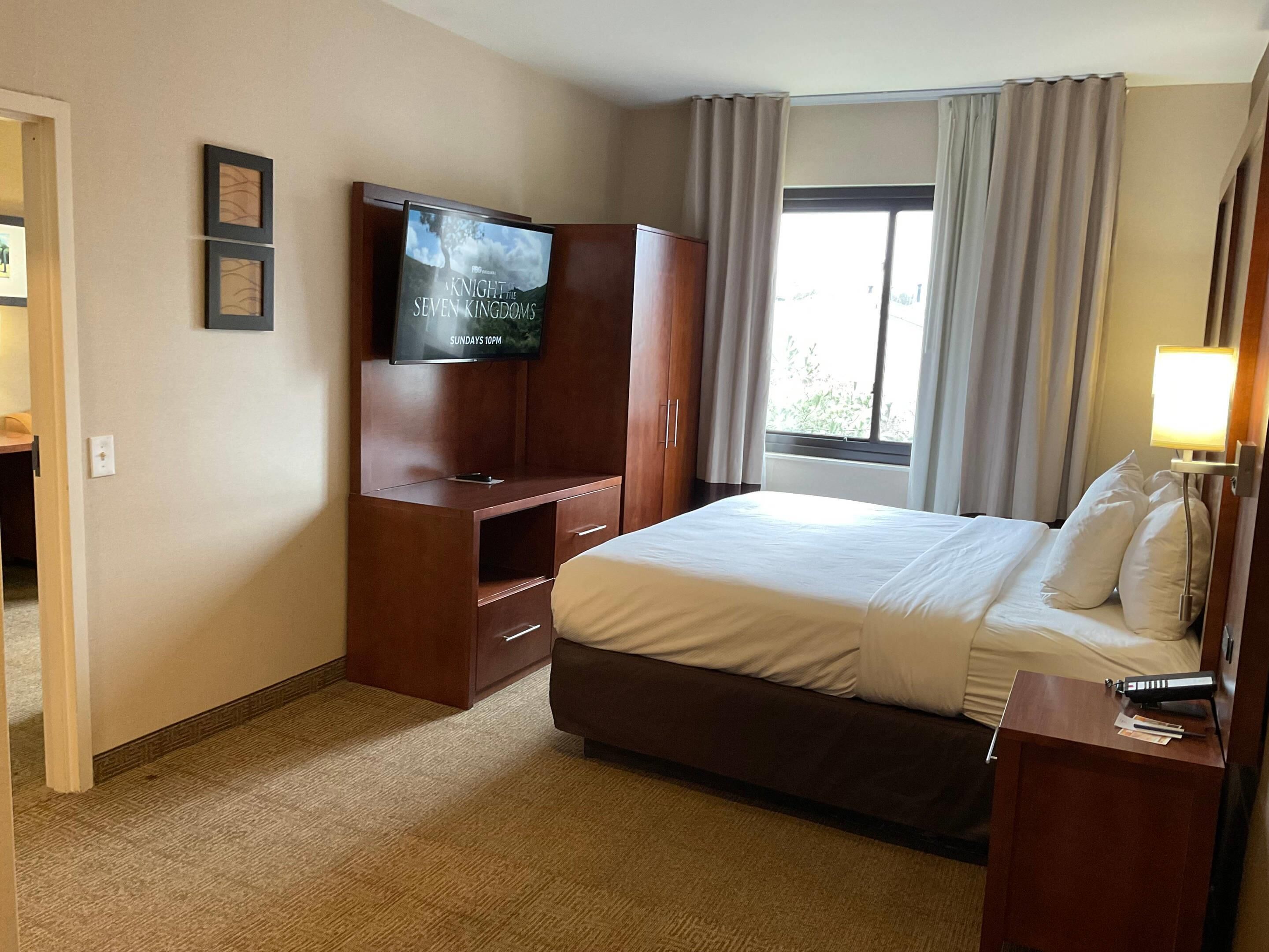 Ein Hotelzimmer mit einem Bett, einem an einem Holzschrank montierten Fernseher, einem Schreibtisch mit einer Lampe und einem Fenster mit Vorhängen.