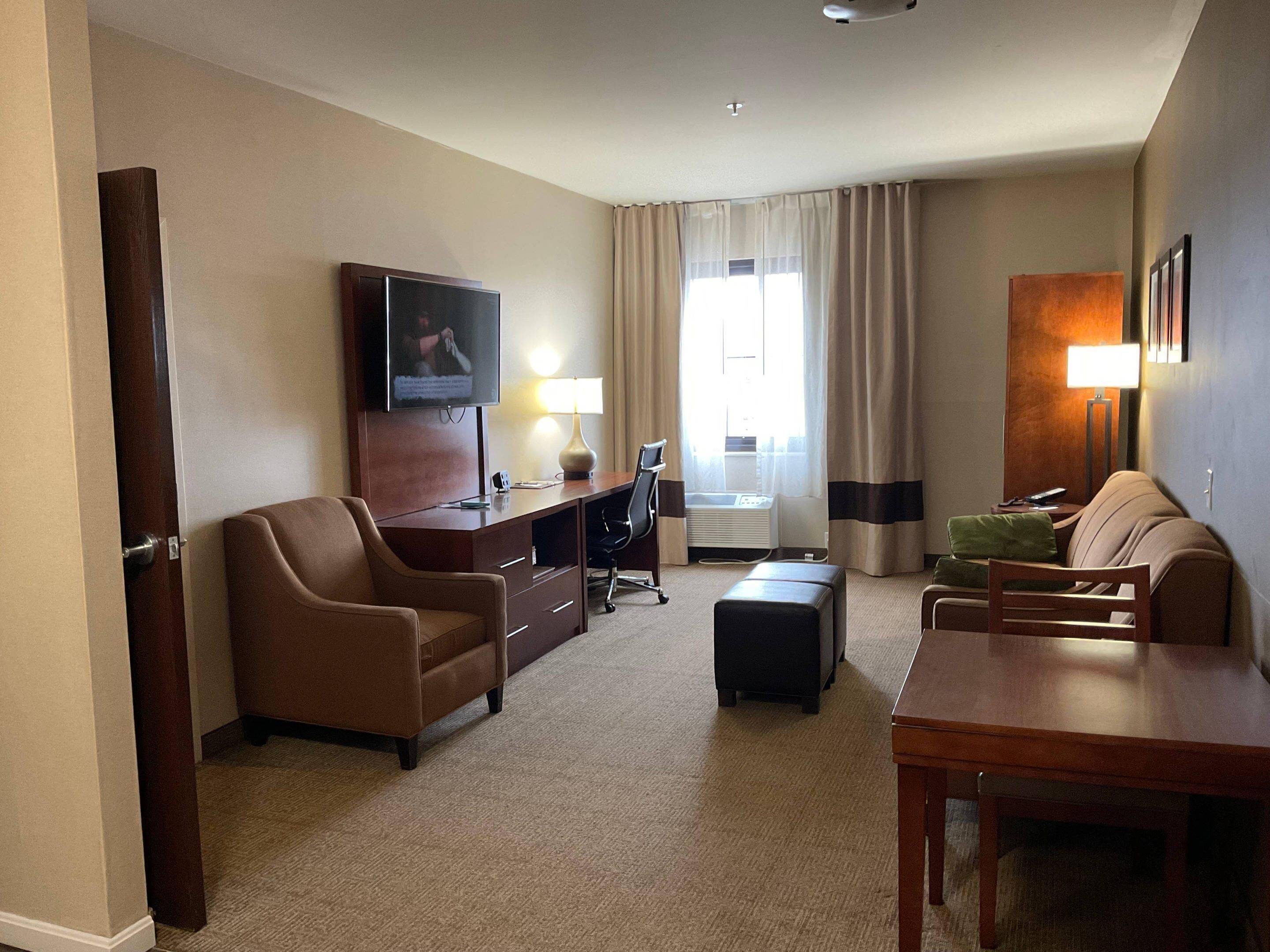 A hotel room with a brown leather sofa, a wooden desk, a television, a lamp, and a window with curtains.
