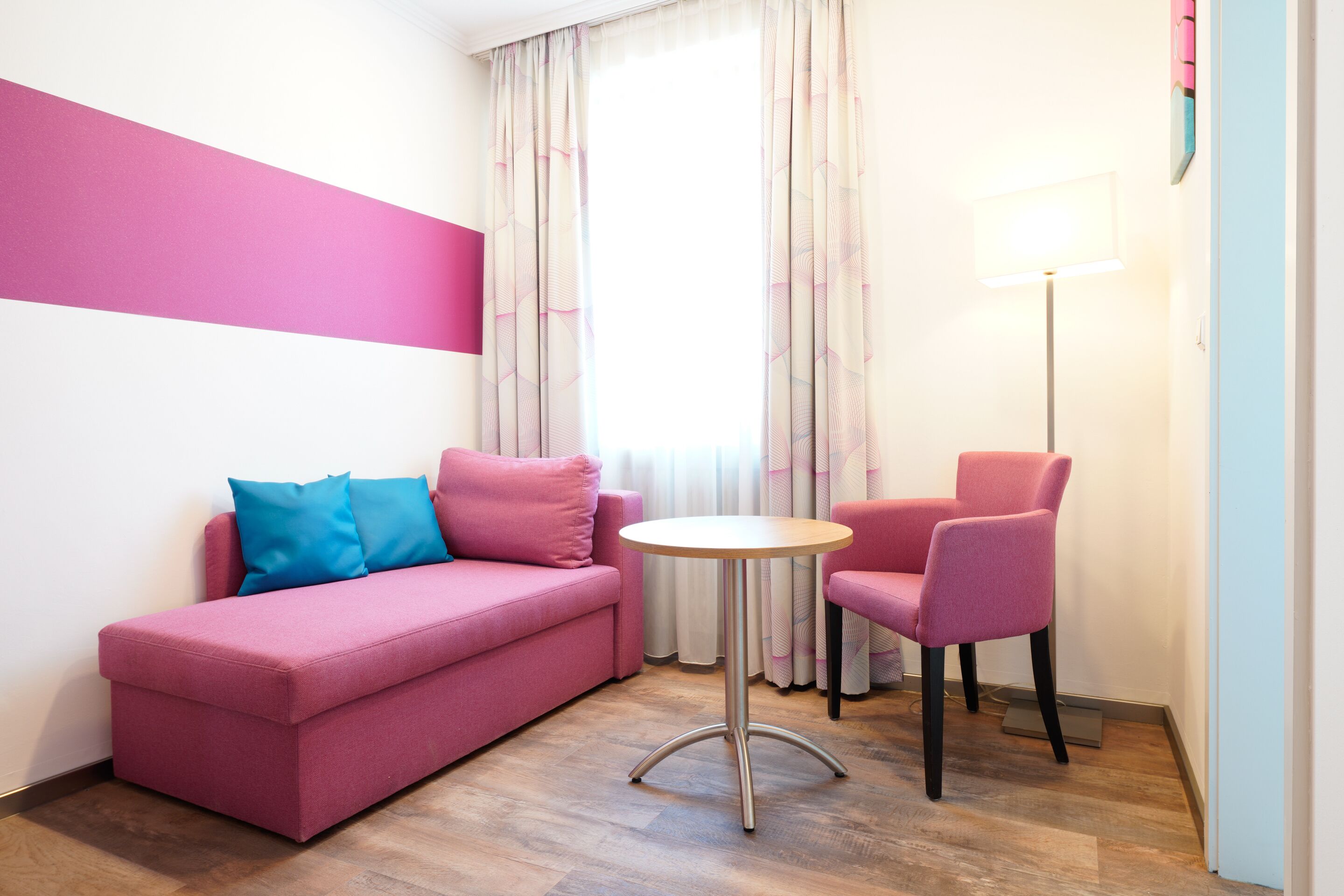 A pink sofa with a blue pillow, a pink chair, a small round table, and a floor lamp in a room with wooden flooring and a window with curtains.