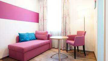 A pink sofa with a blue pillow, a pink chair, a small round table, and a floor lamp in a room with wooden flooring and a window with curtains.