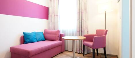 Un sofá rosa con un cojín azul, una silla rosa, una mesita redonda pequeña y una lámpara de pie en una habitación con suelo de madera y una ventana con cortinas.