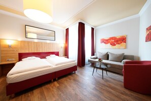 A modern hotel room with a double bed, a sofa, a red armchair, a small table, and a painting on the wall. - Hotel Bayerischer Hof (Bayreuth)