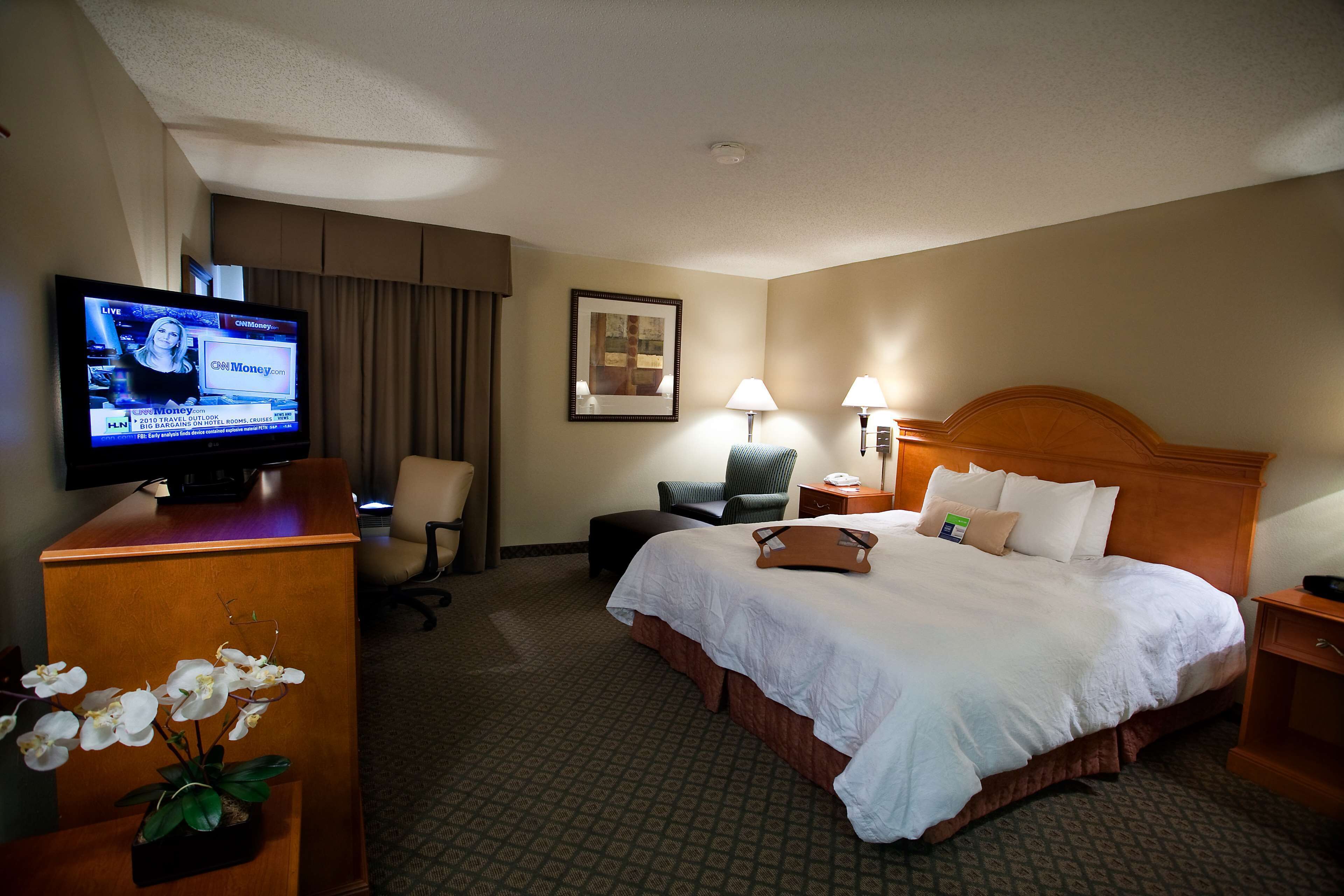 A hotel room with a bed, a desk with a television, a chair, a lamp, and a small table.