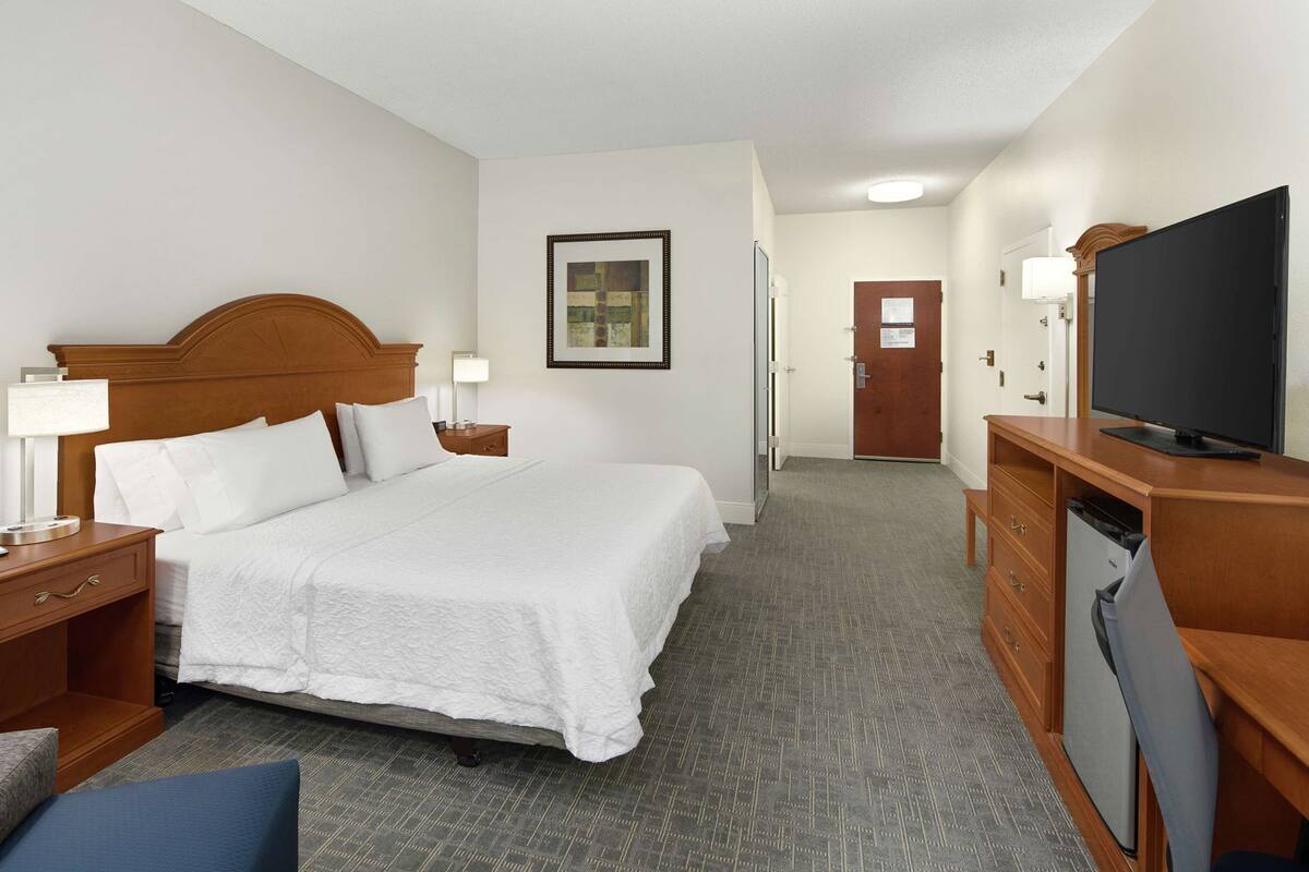 A hotel room with a large bed, a desk with a television, a chair, and a door leading to another room.