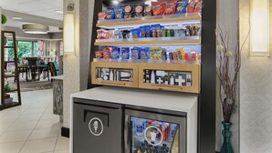 Eine Snackauslage mit Chips und anderen Snacks, ein Kühlschrank mit Getränkeausgabe und eine Vase mit dekorativen Zweigen.