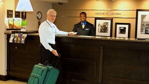 Ein Hotelrezeptionstresen mit einem Mann im Anzug, einem weiteren Mann mit weißem Hemd, einem grünen Koffer und gerahmten Bildern an der Wand.