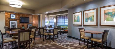 A hotel dining area with multiple tables and chairs, a flat-screen TV mounted on a textured wall, and framed artwork on the walls.