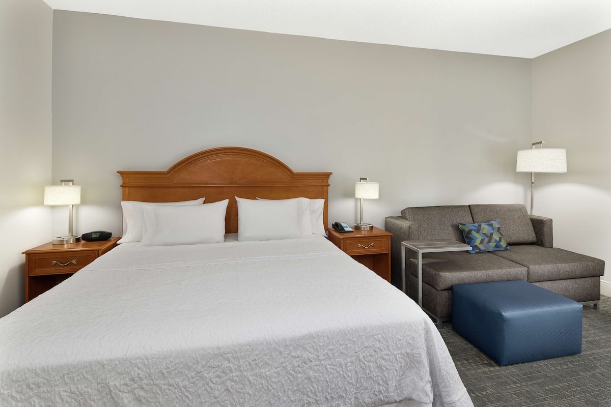 A hotel room with a large bed, a sofa, a nightstand with a lamp, and a small table.