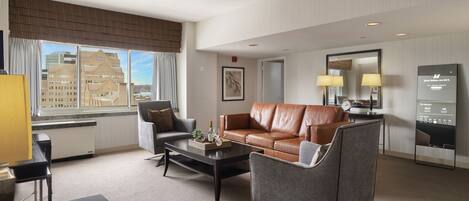 A modern hotel room with a brown leather sofa, a grey armchair, a coffee table, and a large window offering a city view.