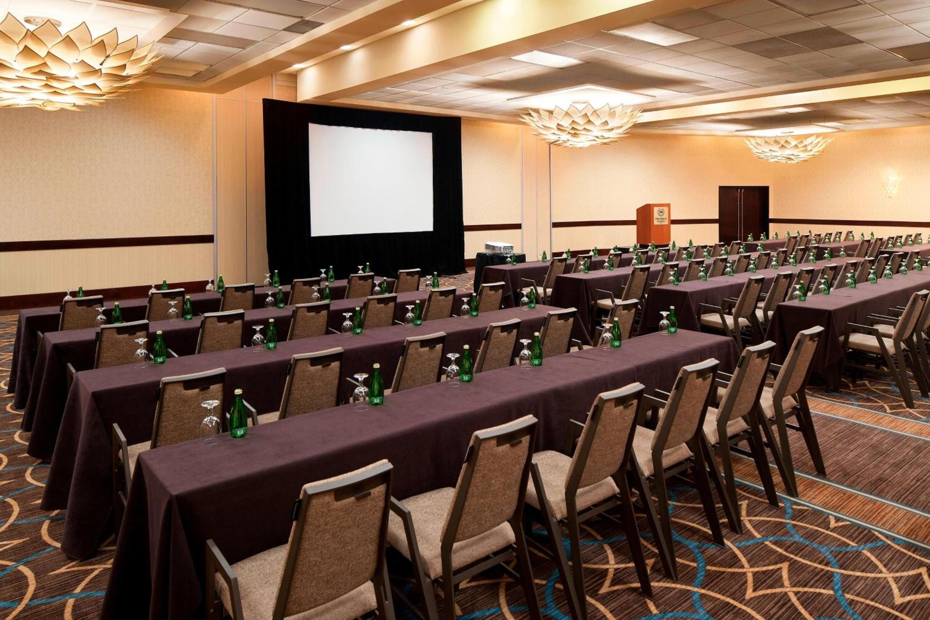 A conference room with rows of tables and chairs, a projector screen, and a podium.