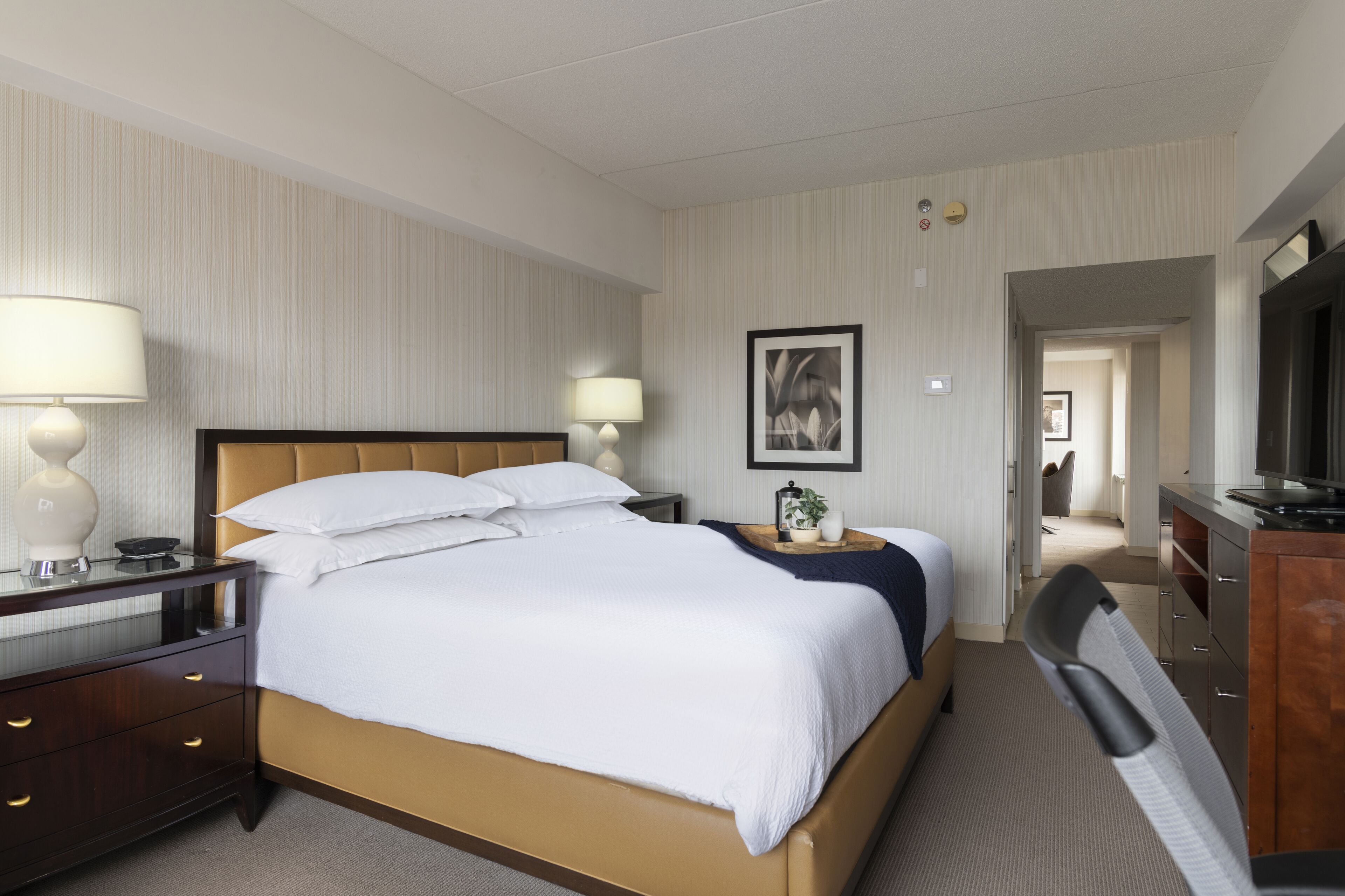 A hotel room with a bed, bedside lamps, a desk, a chair, a television, and a framed picture on the wall.