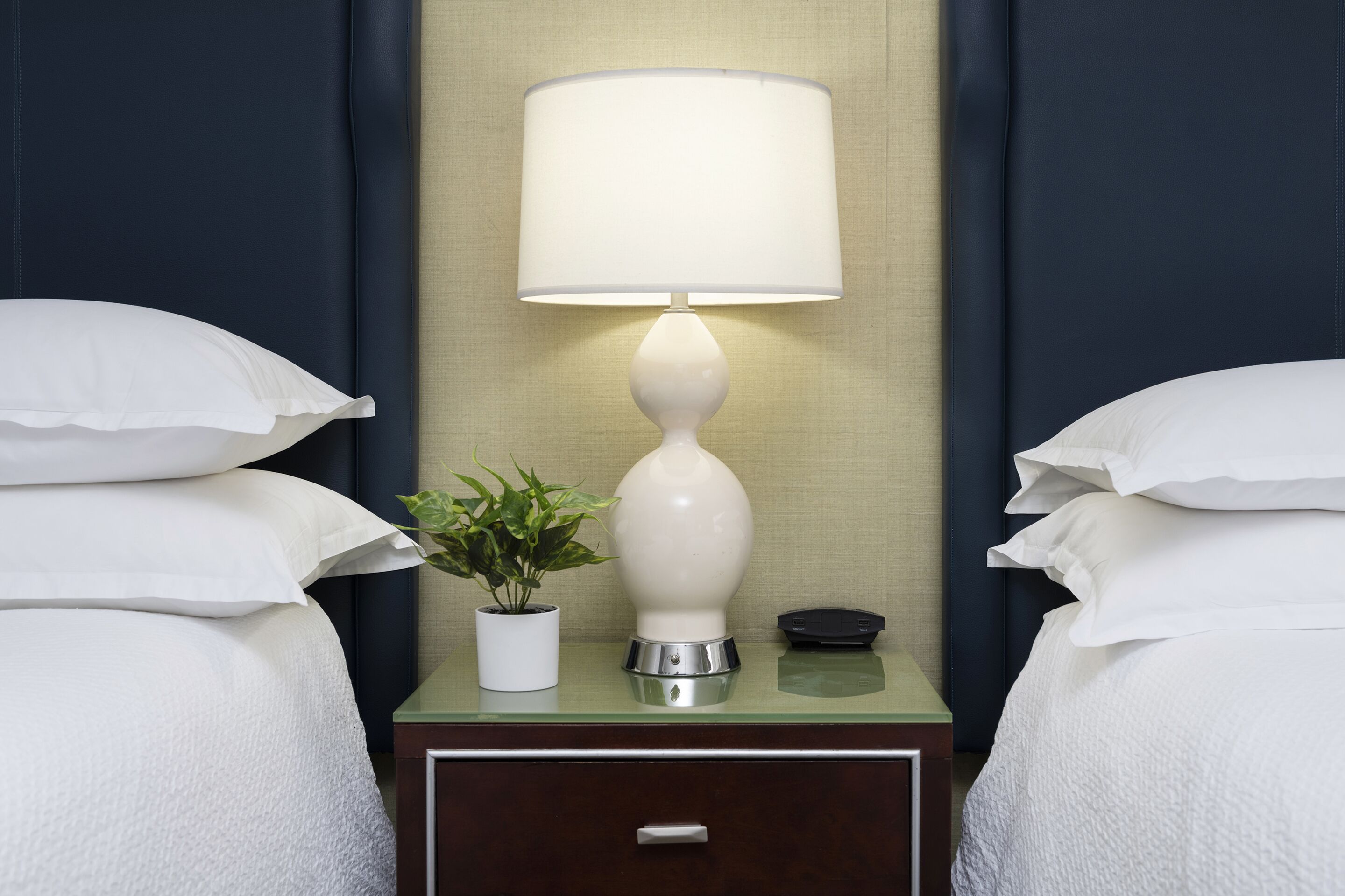A neatly made bed with white bedding, a bedside table with a lamp, a small potted plant, and a bedside clock.