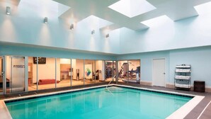 An indoor swimming pool with clear glass walls, a ceiling with skylights, and a visible fitness center in the background.