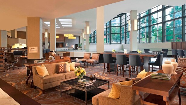 A spacious hotel lobby with a modern design, featuring a mix of seating areas, a dining area, and a view through large windows.