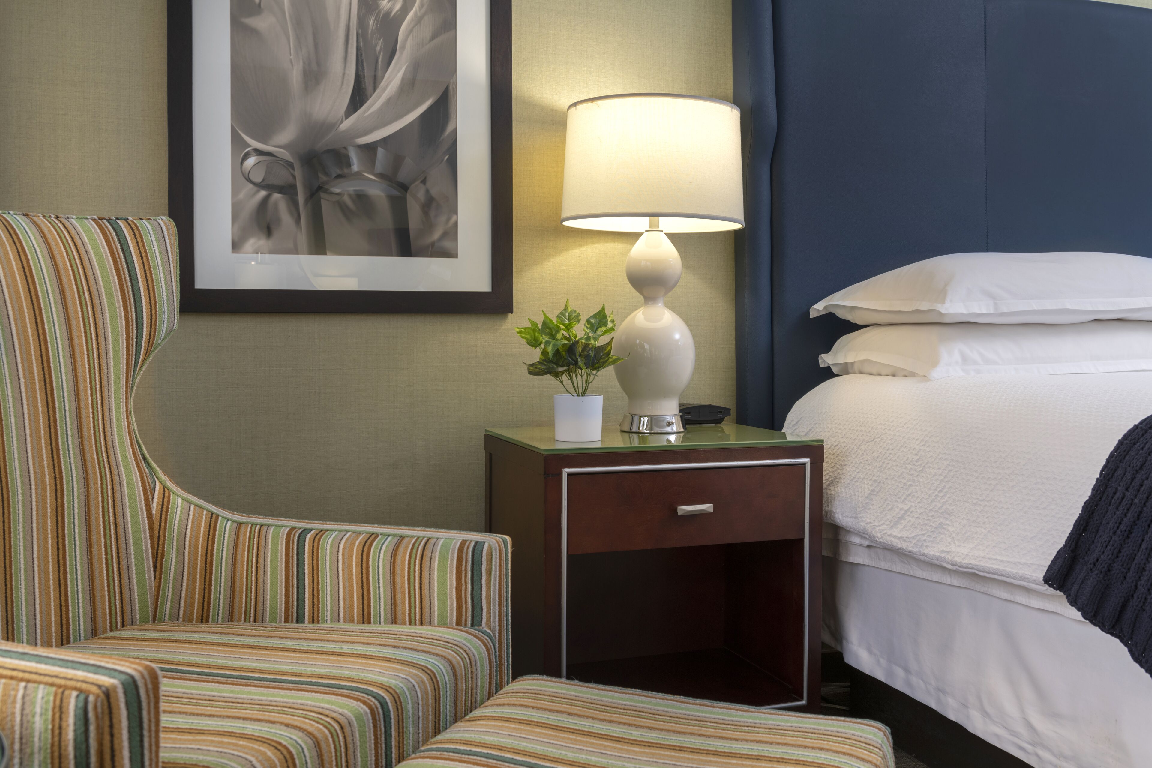 Une chambre d’hôtel avec un fauteuil à rayures, une table de chevet avec une lampe et une petite plante, et un lit bien fait avec du linge de lit blanc.