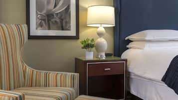 Une chambre d’hôtel avec un fauteuil à rayures, une table de chevet avec une lampe et une petite plante, et un lit bien fait avec du linge de lit blanc.