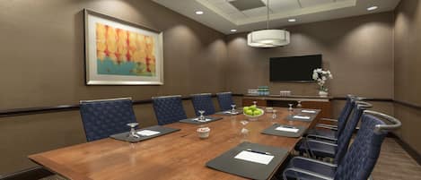 A conference room with a long table, blue chairs, a framed abstract painting, a flat-screen TV, and a vase with flowers.