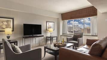 A modern hotel room with a flat-screen TV, a sofa, a chair, a coffee table, and a window with a city view.