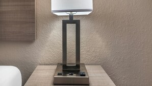 A modern table lamp with a rectangular base and a square lampshade, positioned on a wooden nightstand. - Red Roof Inn Queensbury (Queensbury)