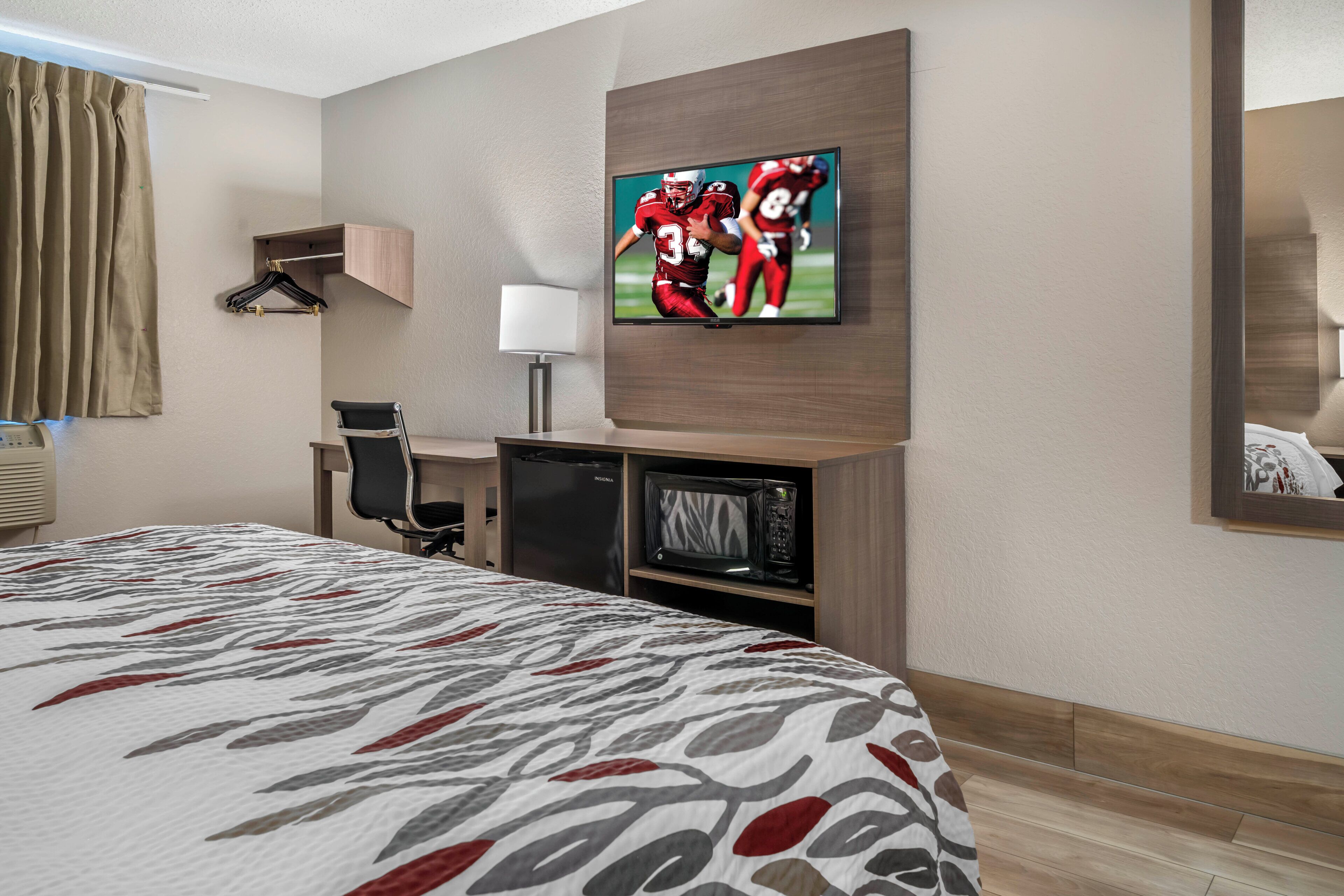 Ein Hotelzimmer mit einem Bett, einem an der Wand befestigten Fernseher, einem Schreibtisch mit Stuhl, einer Mikrowelle und einem kleinen Kühlschrank.