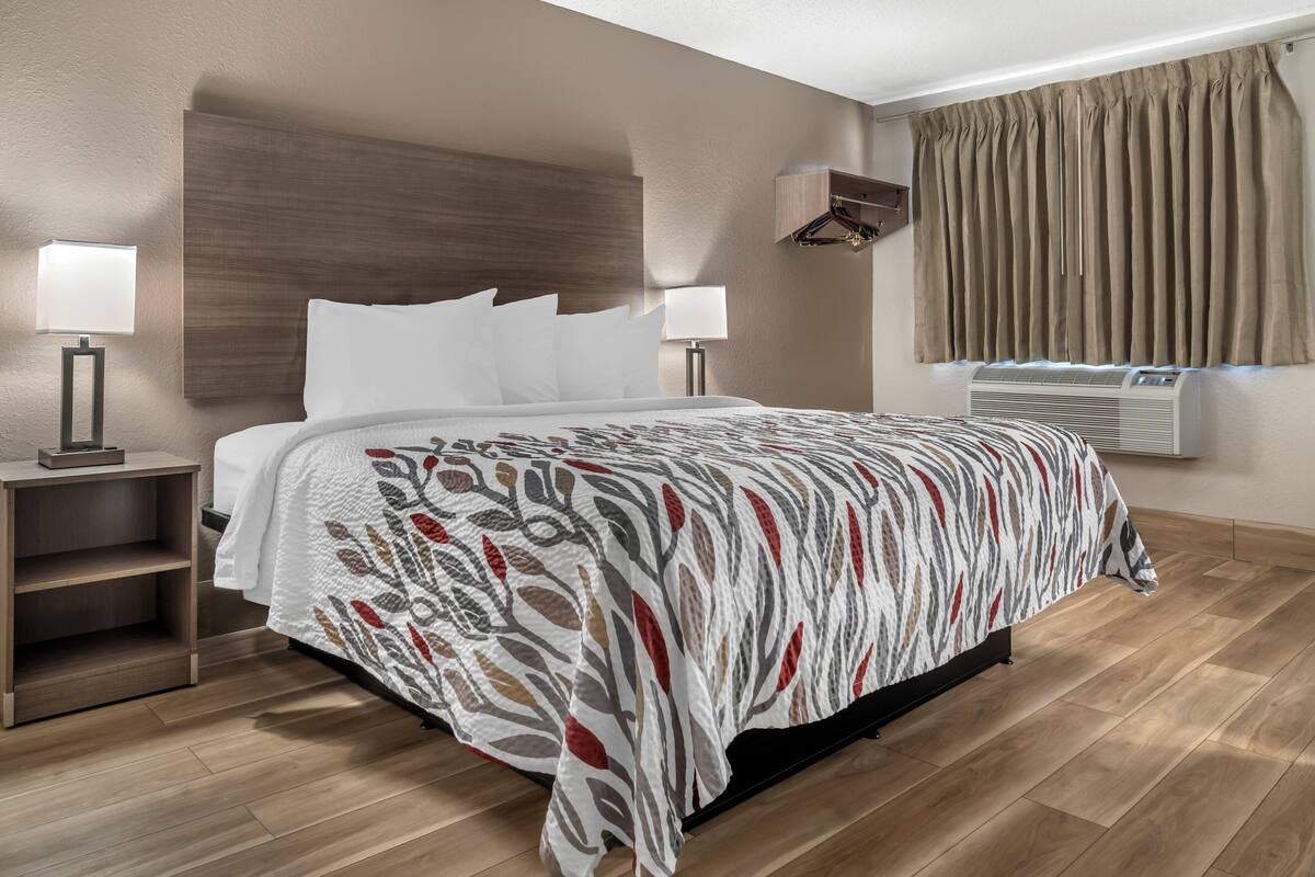 A hotel room with a large bed, two bedside lamps, a wooden headboard, a window with curtains, and an air conditioning unit.