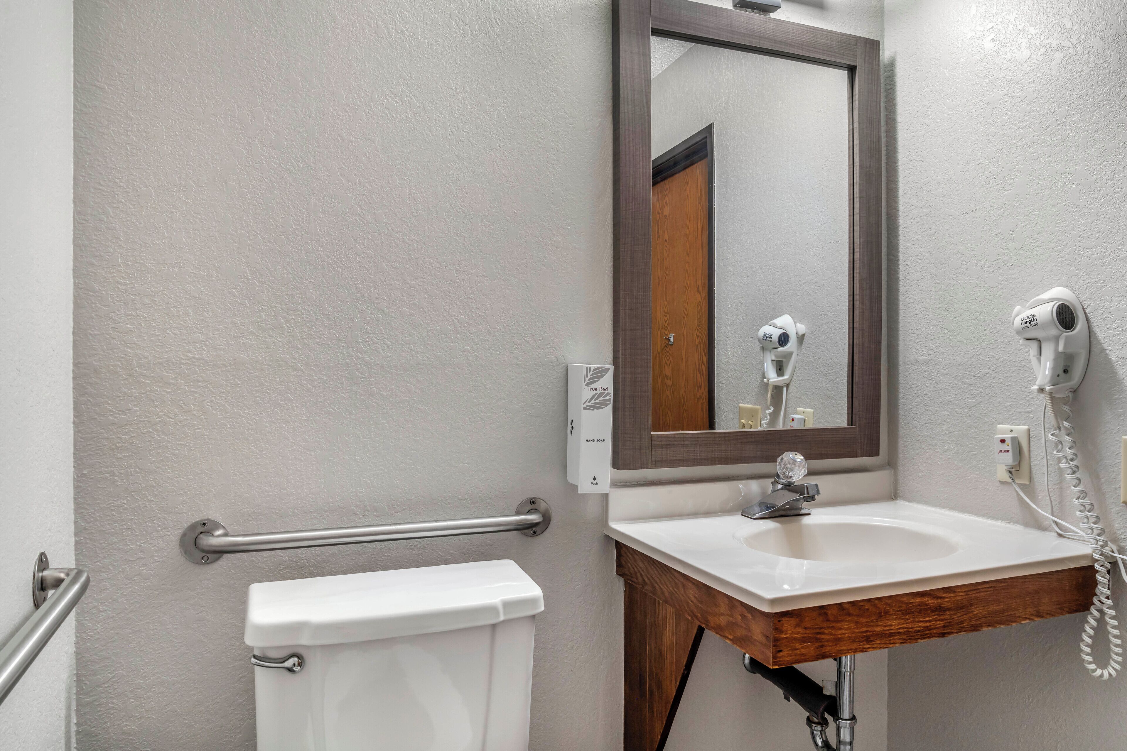 Een badkamer met een wit toilet, een houten wastafelmeubel met een wastafel, een spiegel en een haardroger bevestigd aan de muur.