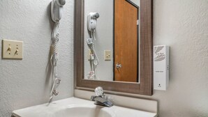 A bathroom with a white sink, a mirror, a hairdryer, and a hand soap dispenser. - Red Roof Inn Queensbury (Queensbury)