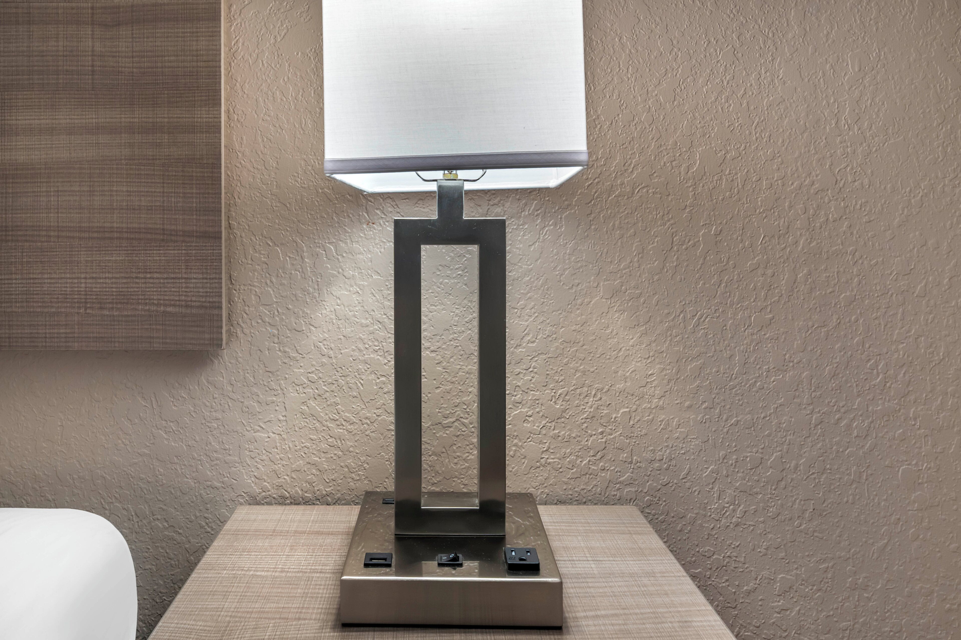 A modern table lamp with a rectangular base and a square lampshade, positioned on a bedside table against a textured wall.