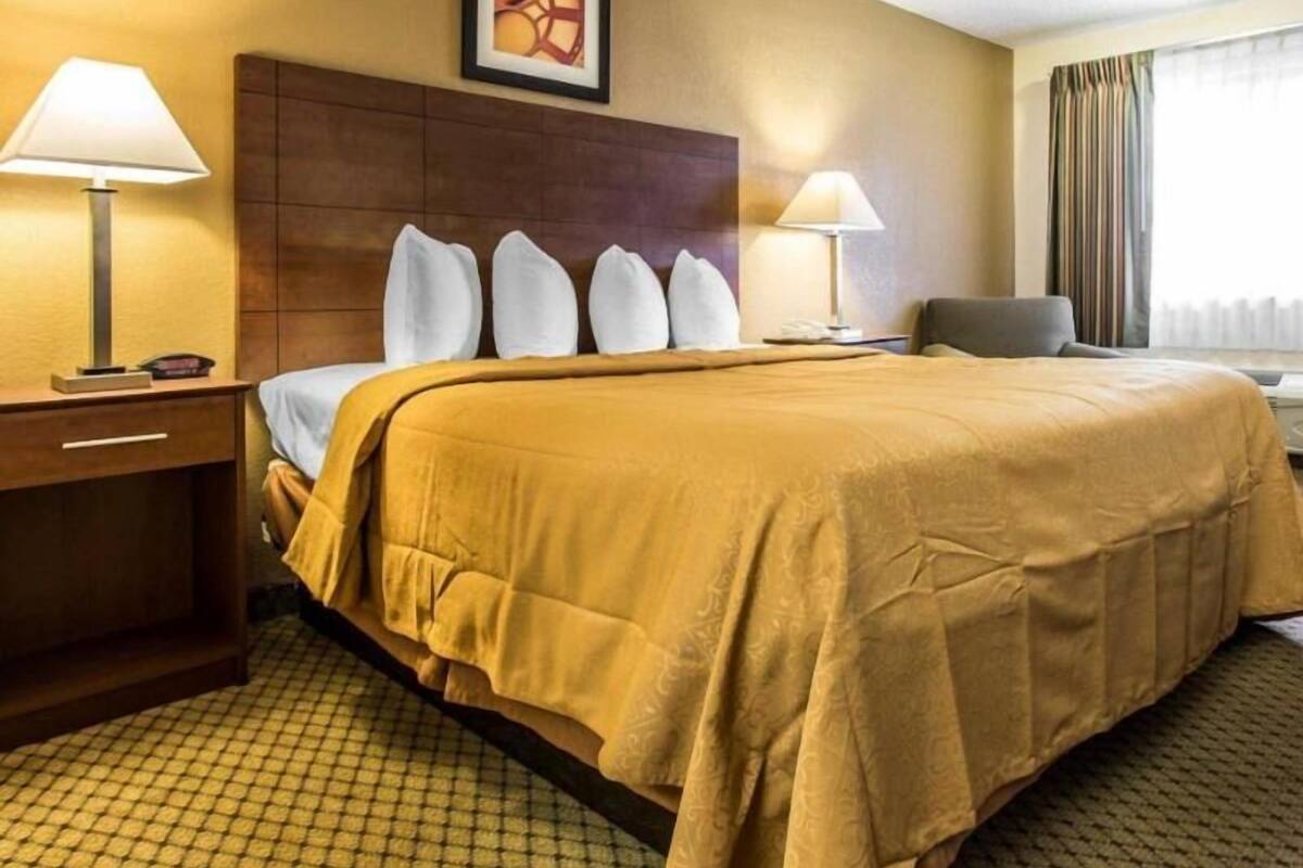 A hotel room with a large bed, two bedside lamps, a headboard, a framed picture on the wall, a chair, and a window with curtains.