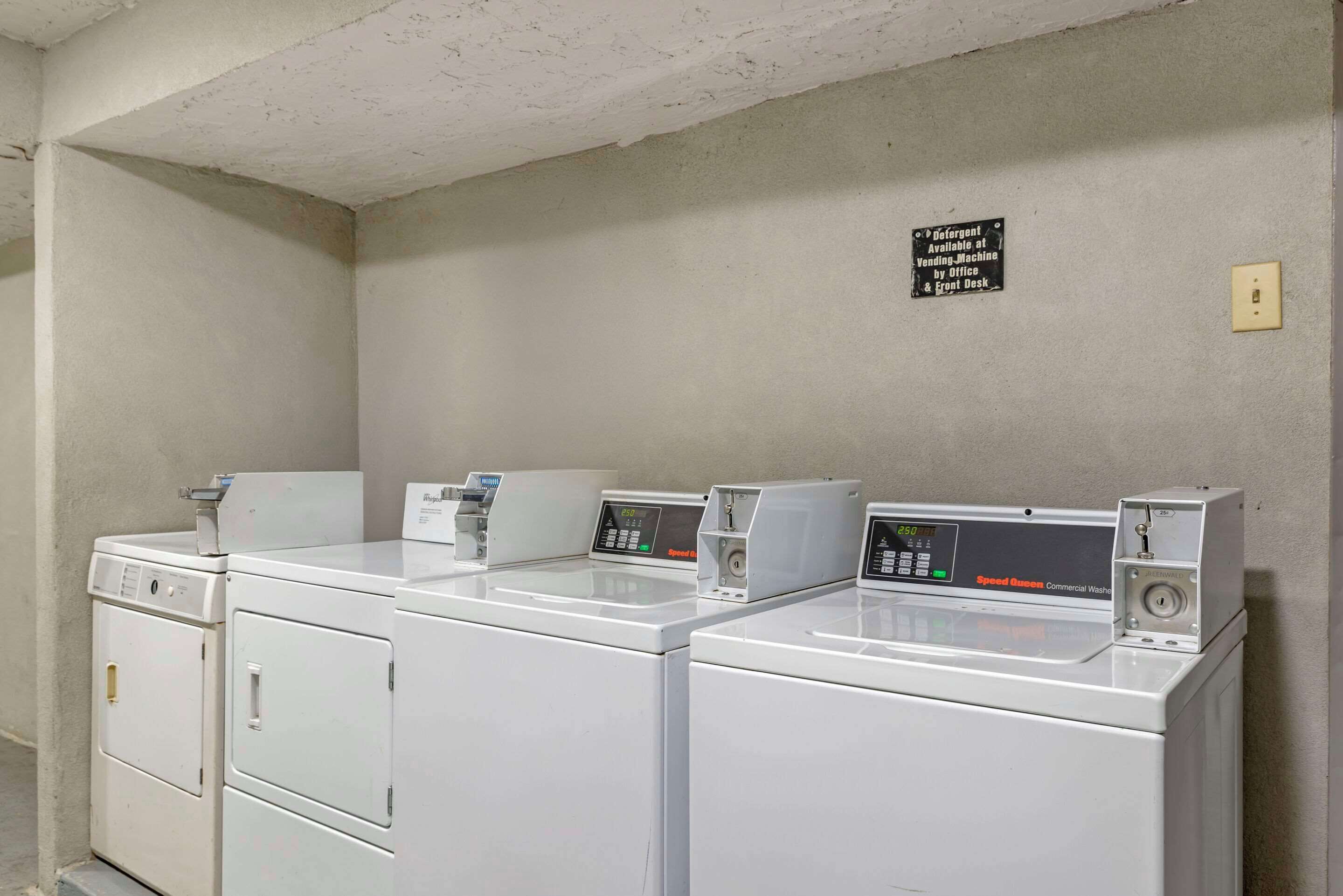 A laundry area with four white commercial washers and dryers, a sign indicating the availability of a vending machine and front desk, and a light switch on the wall.