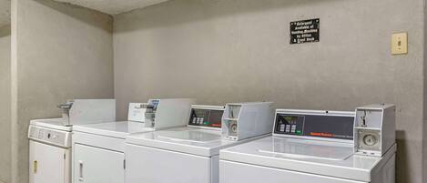 Un área de lavandería con cuatro lavadoras y secadoras comerciales blancas, un cartel que indica la disponibilidad de una máquina expendedora y una recepción, y un interruptor de luz en la pared.