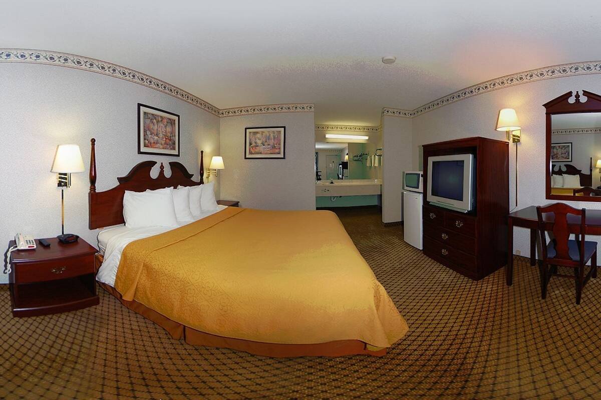 Habitación de hotel con cama, mesita de noche, lámpara, televisión, escritorio, silla y espejo.