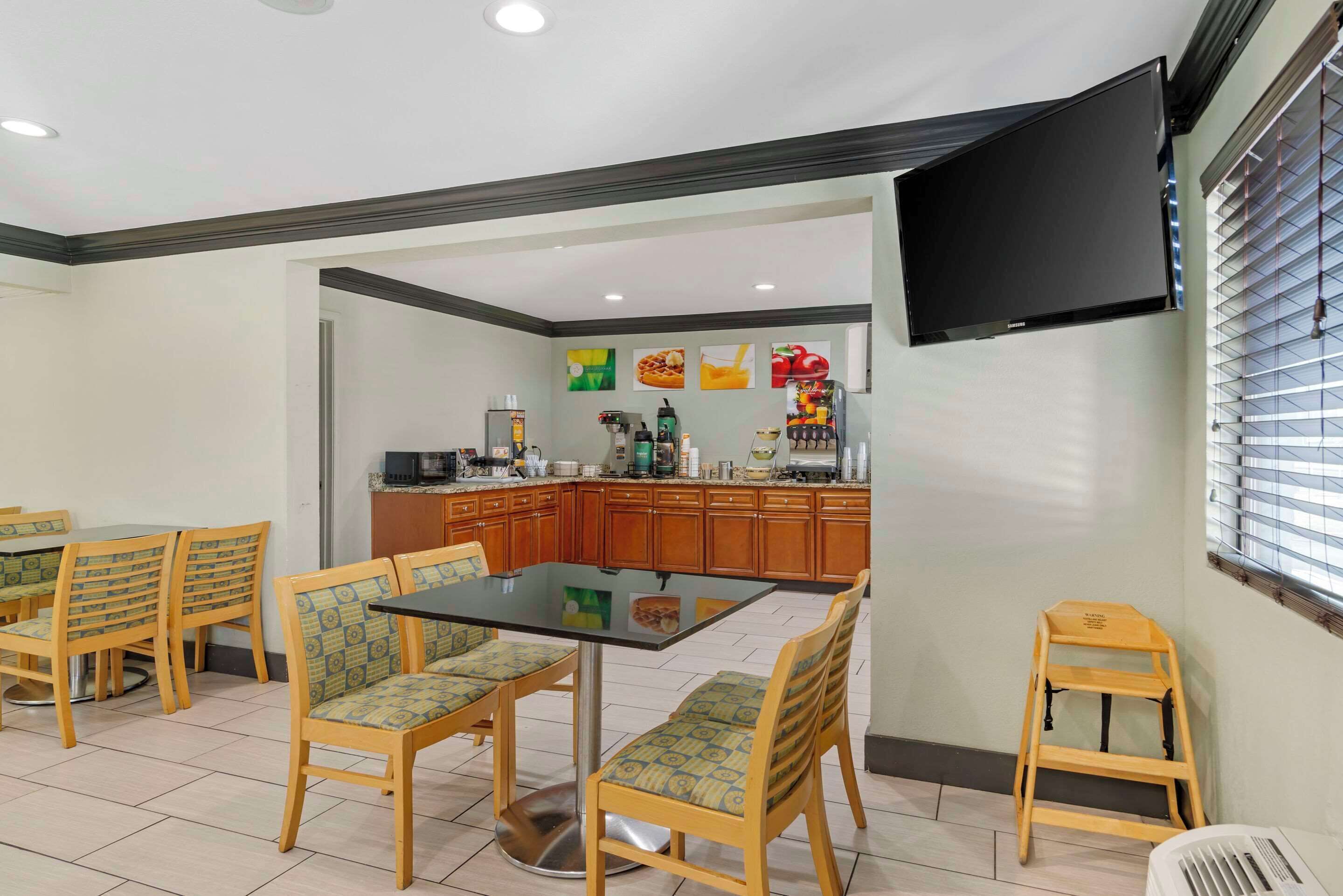 A dining area with a glass table and chairs, a kitchen in the background, and a TV mounted on the wall.