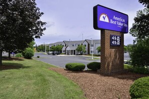 A brick pillar with a sign for Ameris Best Value Inn, a parking lot, and a building with a grey roof. - Americas Best Value Inn East Syracuse (East Syracuse)