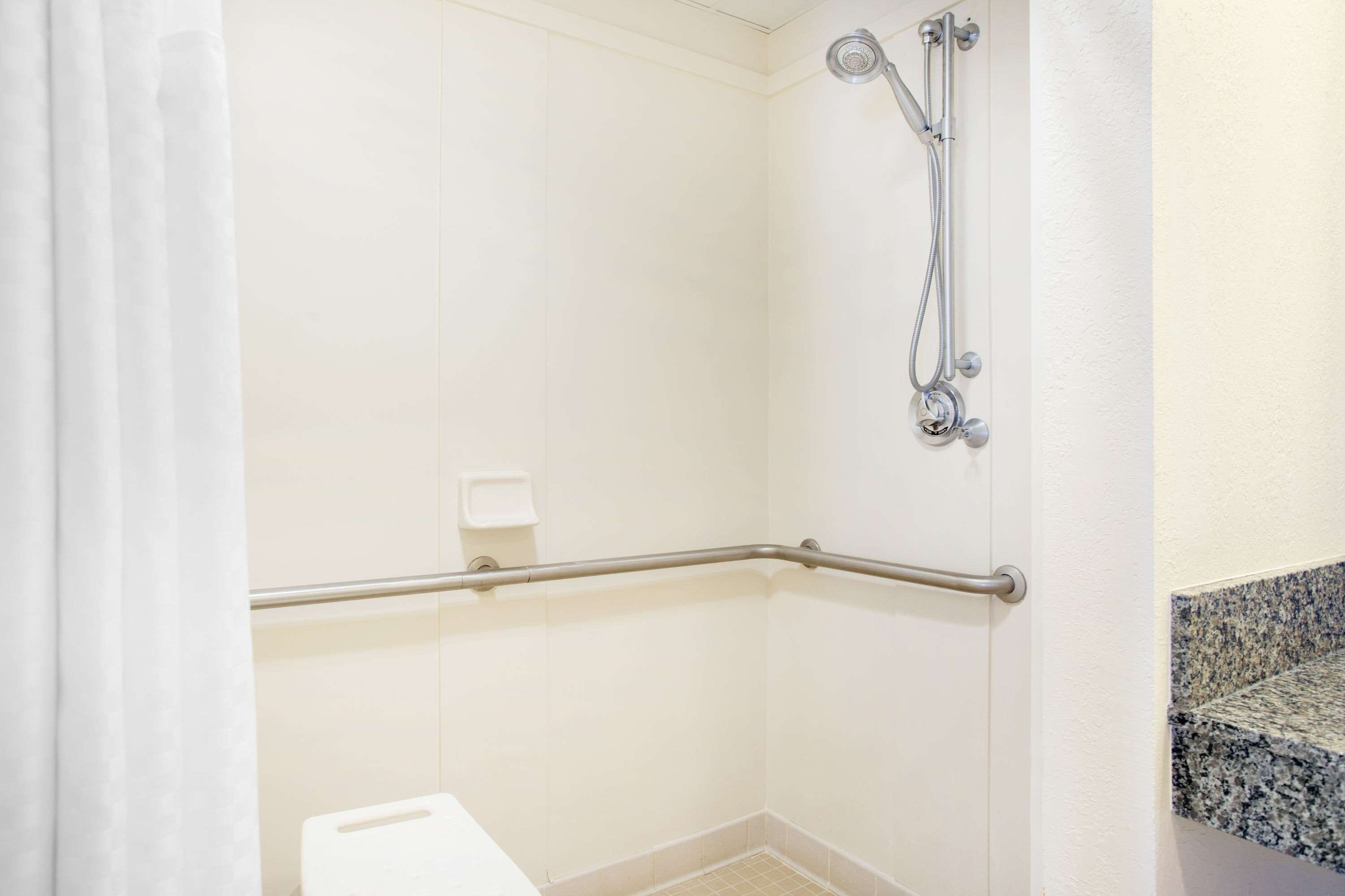 Un bagno con piastrelle bianche, una doccia con soffione manuale, una barra di sostegno e un water bianco.