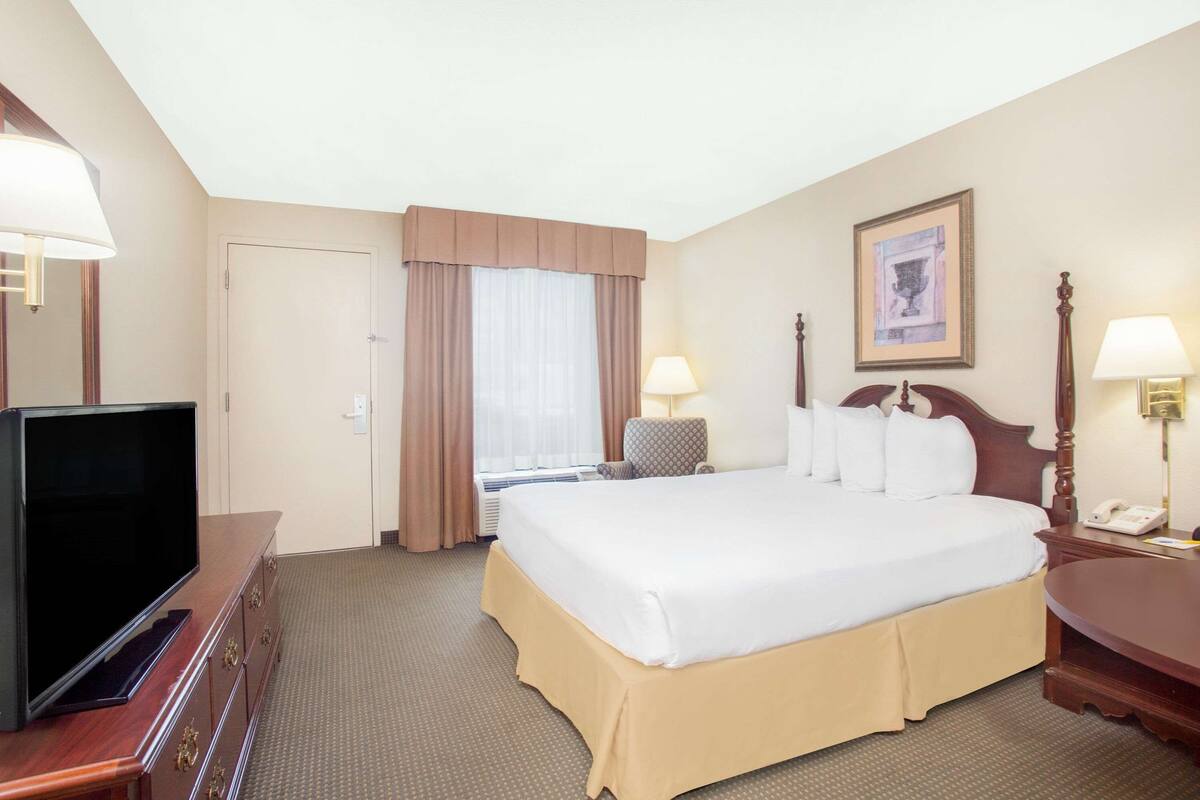 Une chambre d’hôtel avec un grand lit, un téléviseur posé sur une commode, un bureau avec une lampe et une fenêtre avec des rideaux.