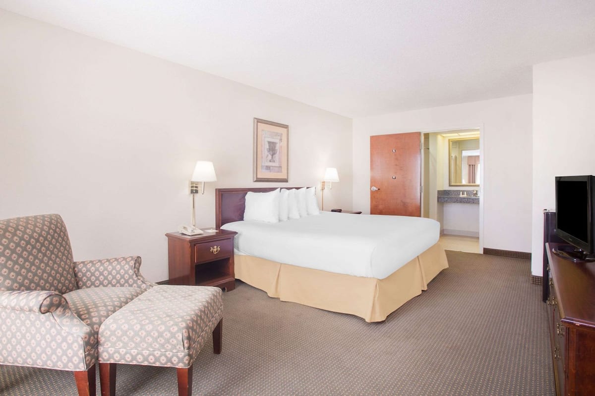 Une chambre d’hôtel avec un lit, des tables de chevet, une chaise, un téléviseur et une salle de bain visible à travers une porte ouverte.