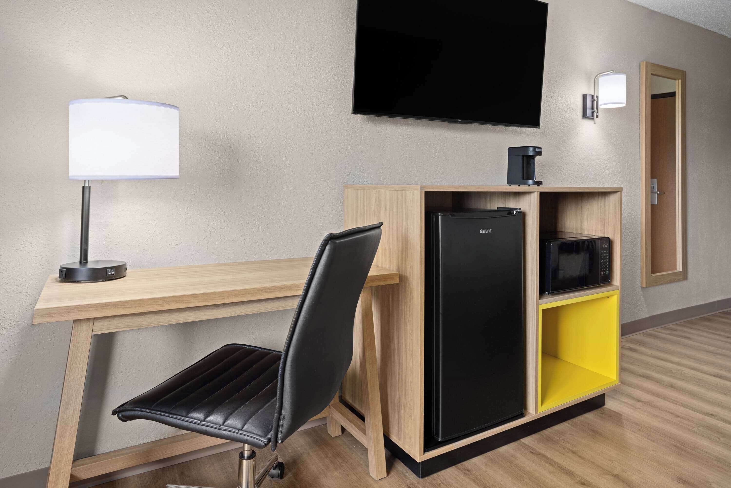 Une chambre d’hôtel équipée d’un bureau, d’une chaise, d’un téléviseur à écran plat, d’une machine à café, d’un réfrigérateur et d’un meuble de rangement jaune.